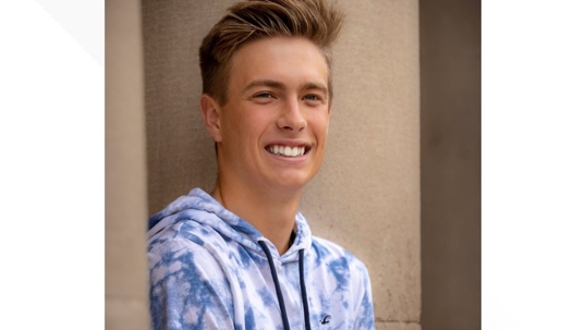 A smiling young man with blond hair, wearing a blue and white hoodie.